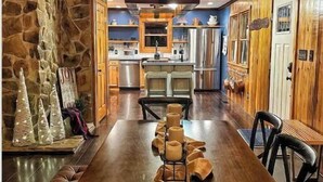 A spacious dining area with a long wooden table, stone wall, and a kitchen in the background.