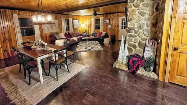 A cozy cabin interior with a dining area, a living room with a stone fireplace, and wooden walls.
