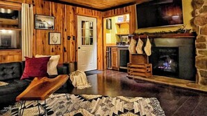 A cozy cabin interior with a wooden ceiling, a fireplace, a stone wall, and a television mounted above it.