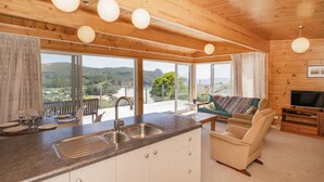 Une cuisine moderne dotée d’une grande fenêtre, un coin repas avec une table dressée pour quatre personnes, et un salon confortable avec un canapé et une télévision.