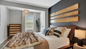 Ein Schlafzimmer mit Bett, Nachttisch, Lampe, Kommode und einem großen Fenster mit Vorhängen.