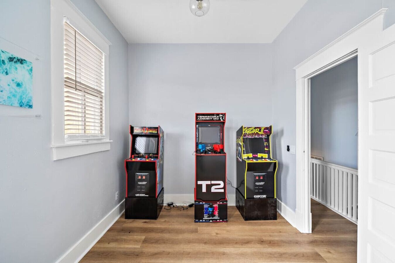 Un pasillo con tres máquinas de videojuegos, una ventana con persianas y una puerta blanca.
