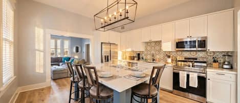 Une cuisine moderne dotée d’un îlot central, de meubles blancs, d’appareils électroménagers en acier inoxydable et d’un luminaire suspendu.