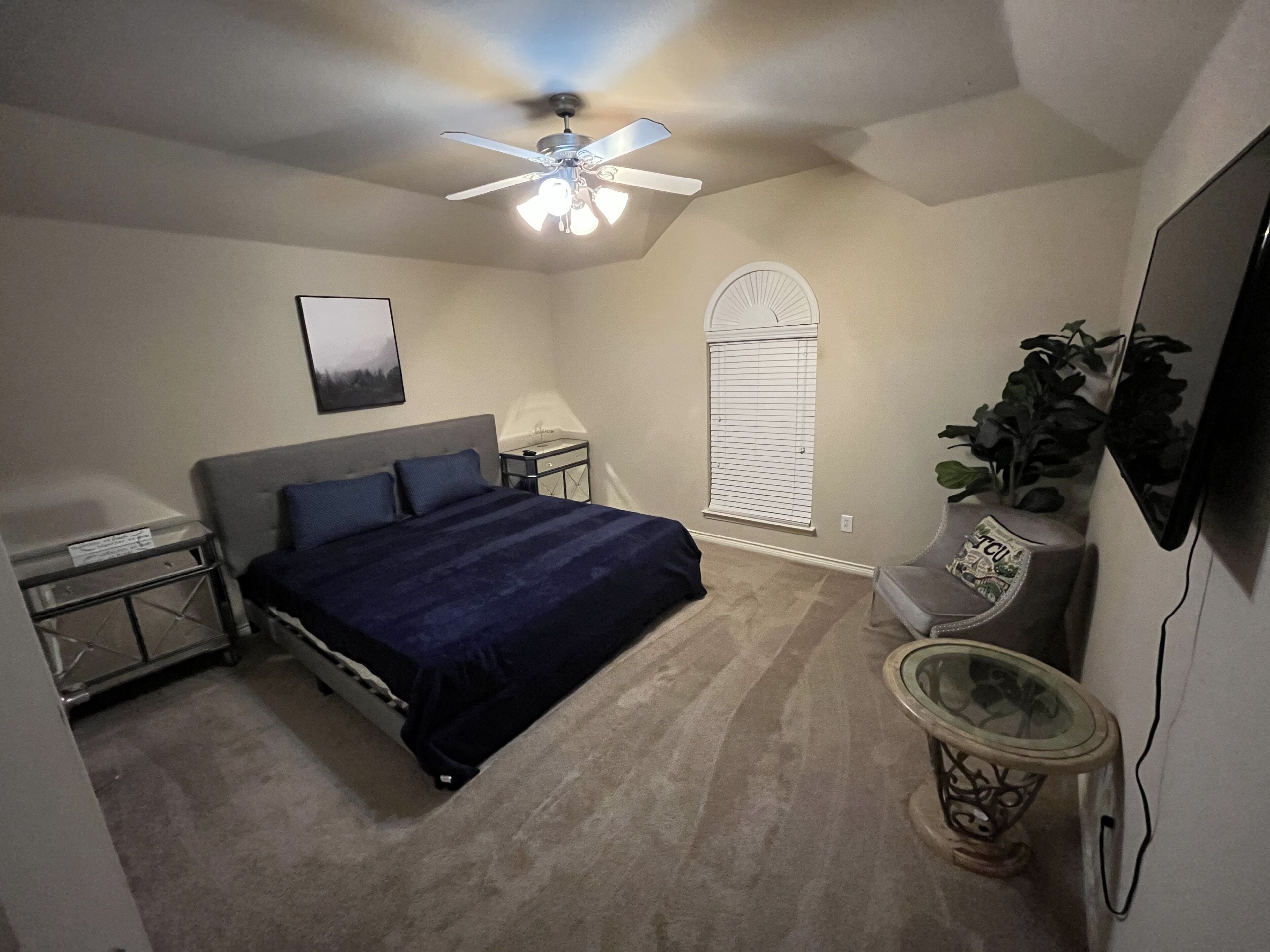 A bedroom with a bed, a ceiling fan, a nightstand, a lamp, a TV, and a window with blinds.