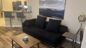 Een woonkamer met een zwarte bank, twee barkrukken, een houten salontafel met een autootje en een schilderij van een stadsgezicht, met op de achtergrond een keuken.