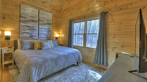 Een slaapkamer in een houten hut met een bed, een groot raam en een bevestigde televisie.