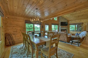A wooden cabin interior with a dining area, fireplace, and large windows.