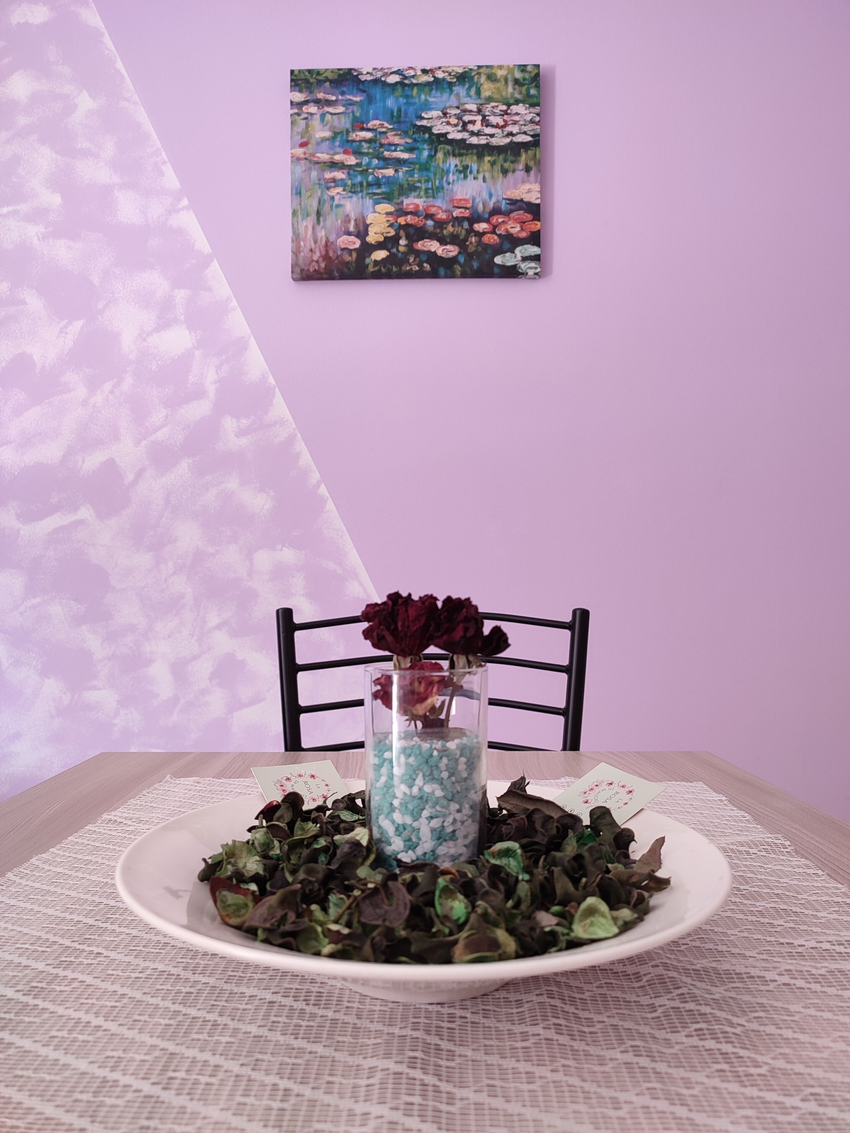 A dining table with a white plate of salad, a vase with dried flowers, and a painting on the wall.