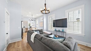 A modern living room with a grey sectional sofa, a flat-screen TV mounted on the wall, and a chandelier. - Charming Home Near Downtown with Pool & Fire Pit (Galveston)