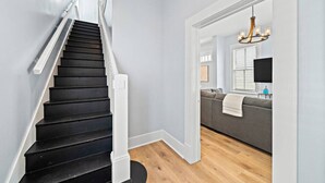 A staircase with black steps and white handrails, leading to a room with a grey sofa and a chandelier. - Charming Home Near Downtown with Pool & Fire Pit (Galveston)