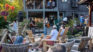 An outdoor seating area with wooden chairs and tables, a musician performing, and a building with a balcony in the background.