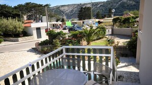 Un balcon avec une table ronde et des chaises, donnant sur une rue où sont garées des voitures et sur un immeuble avec un toit rouge.