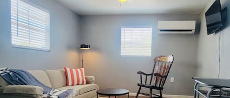 A compact living room with a ceiling fan, a sofa with a throw blanket, a rocking chair, a small table, and a colorful rug.