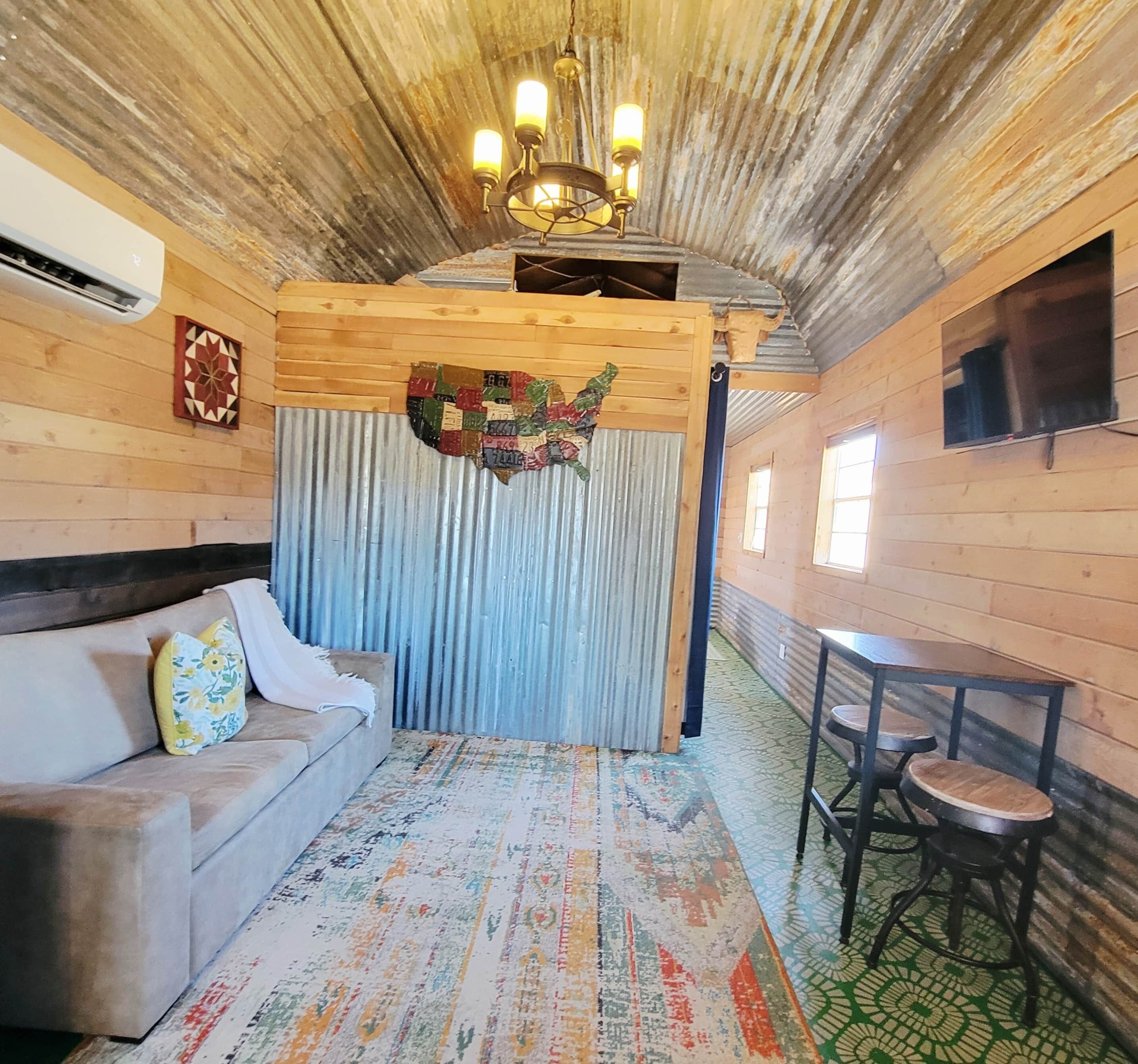 A cozy room with a wooden ceiling, a sofa with floral cushions, a patterned rug, a small table with stools, and a wall-mounted TV.