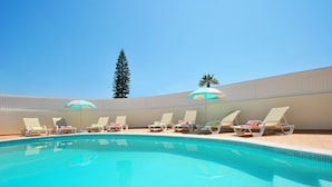 Une piscine entourée de chaises longues et de parasols, le tout délimité par un petit muret et surplombé d’un grand arbre.