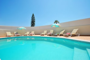 Una piscina con sillas de descanso y sombrillas, rodeada por un muro bajo y un árbol alto.