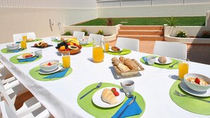 Un espace de restauration en plein air avec des tables préparées pour le petit-déjeuner, où l’on propose une variété de fruits, de viennoiseries et de boissons.