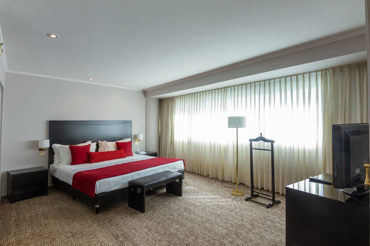Une chambre d’hôtel avec un lit, des tables de chevet, un bureau avec un ordinateur, une lampe et une fenêtre avec des rideaux.