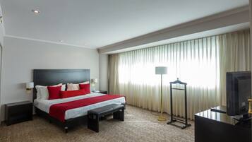 Habitación de hotel con cama, mesitas de noche, escritorio con computadora, lámpara y ventana con cortinas.