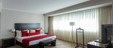A hotel room with a bed, bedside tables, a desk with a computer, a lamp, and a window with curtains.