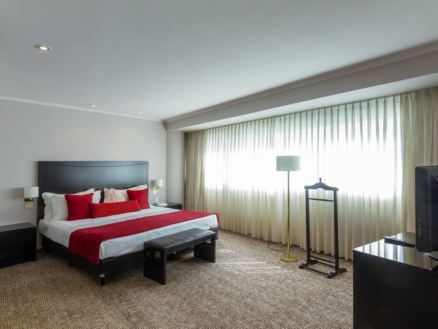 Habitación de hotel con cama, mesitas de noche, escritorio con computadora, lámpara y ventana con cortinas.