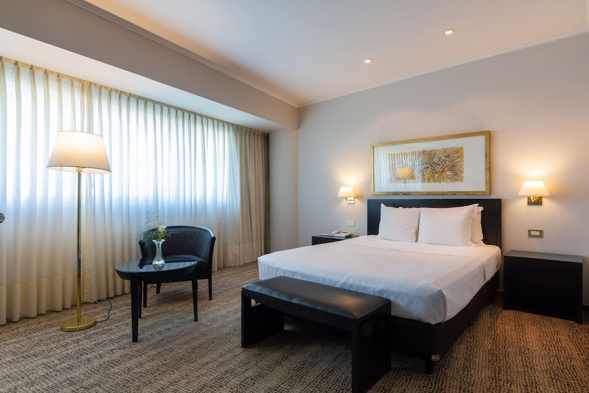 Une chambre d’hôtel avec un grand lit, des lampes de chevet, une petite table avec un vase, un banc et une grande fenêtre avec des rideaux.
