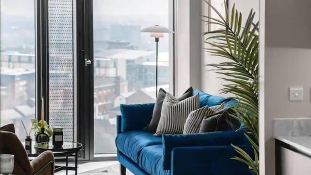 Een moderne woonkamer met een blauwe bank, een potplant en uitzicht op de stad door een groot raam.