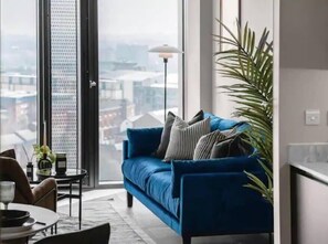 Ein modernes Wohnzimmer mit einem blauen Sofa, einer Topfpflanze und Blick auf die Stadt durch ein großes Fenster.