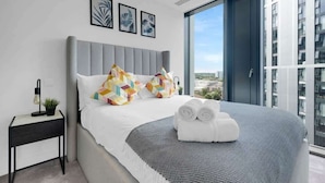 Moderni hotellihuone, jossa on suuri sänky, pieni sivupöytä ja näkymä kaupunkiin suurista ikkunoista.