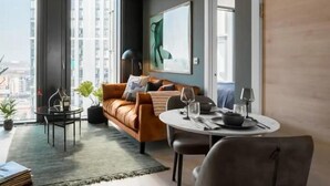 A modern living room with a leather sofa, a dining table set for two, and a large window offering a city view.