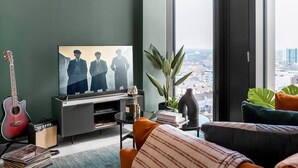 A modern living room with a large flat-screen TV, a guitar, and a city view through large windows. - The Mercian 1 XVIII- 1BDR |STUNNING CITY SKYLINE VIEWS! 24-Hour GYM | CINEMA room (West Midlands)
