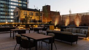 An outdoor seating area with tables and sofas, surrounded by buildings. - The Mercian 2 I - 1BDR |STUNNING CITY SKYLINE VIEWS! 24-Hour GYM | CINEMA room (West Midlands)