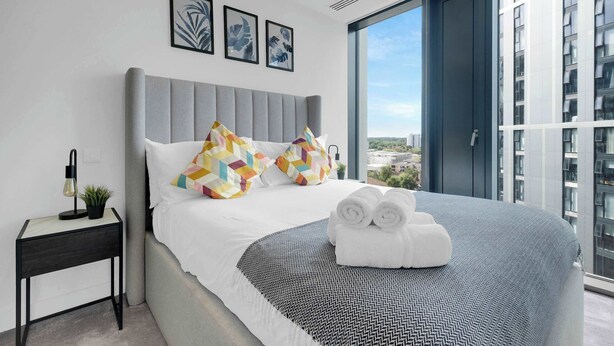 A modern hotel room with a large bed, grey headboard, and a city view through the window. - The Mercian 2 I - 1BDR |STUNNING CITY SKYLINE VIEWS! 24-Hour GYM | CINEMA room (West Midlands)