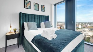 A modern hotel room with a large bed, a nightstand with a lamp, and a city view through the window. - The Mercian 2 VIII - 1BDR | STUNNING CITY SKYLINE VIEWS! 24-Hour GYM | CINEMA room (West Midlands)