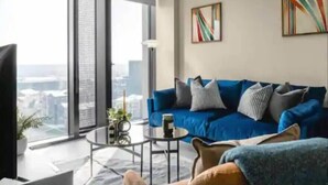 A modern living room with a blue sofa, a coffee table, and a large window offering a city view. - The Mercian 2 VIII - 1BDR | STUNNING CITY SKYLINE VIEWS! 24-Hour GYM | CINEMA room (West Midlands)