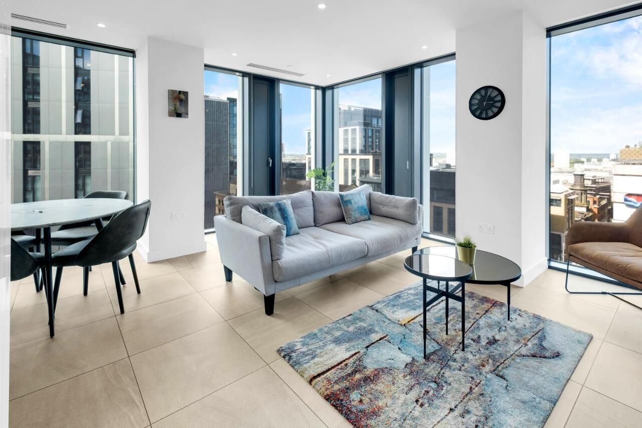 A modern living room with a grey sofa, a dining table with chairs, a coffee table, and a large window offering a city view.