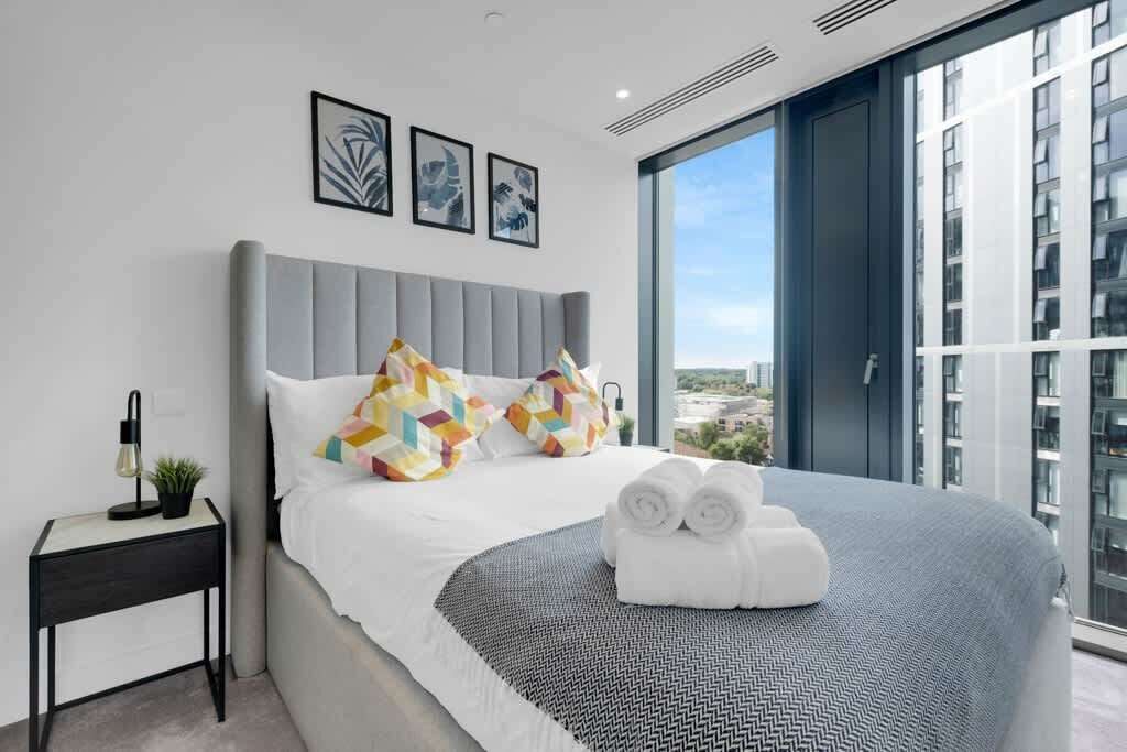 A modern hotel room with a large bed, a small side table, and a view of the city through large windows.