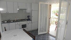 A compact kitchen with white cabinets, a stove, and a sink. A dining table with a white tablecloth is in the foreground. The room has a large sliding glass door leading to an outdoor area with a view of a garden.