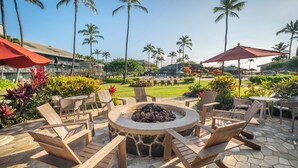 A circular fire pit with wooden chairs and tables, surrounded by a stone patio, palm trees, and a view of a golf course. - Oceanfront Resort, 2 QUEEN beds, water views, walk-out access to pools/ocean (Lihue)