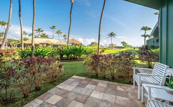 A patio area with white chairs, a checkered floor, and a view of a golf course with palm trees and thatched-roof structures.