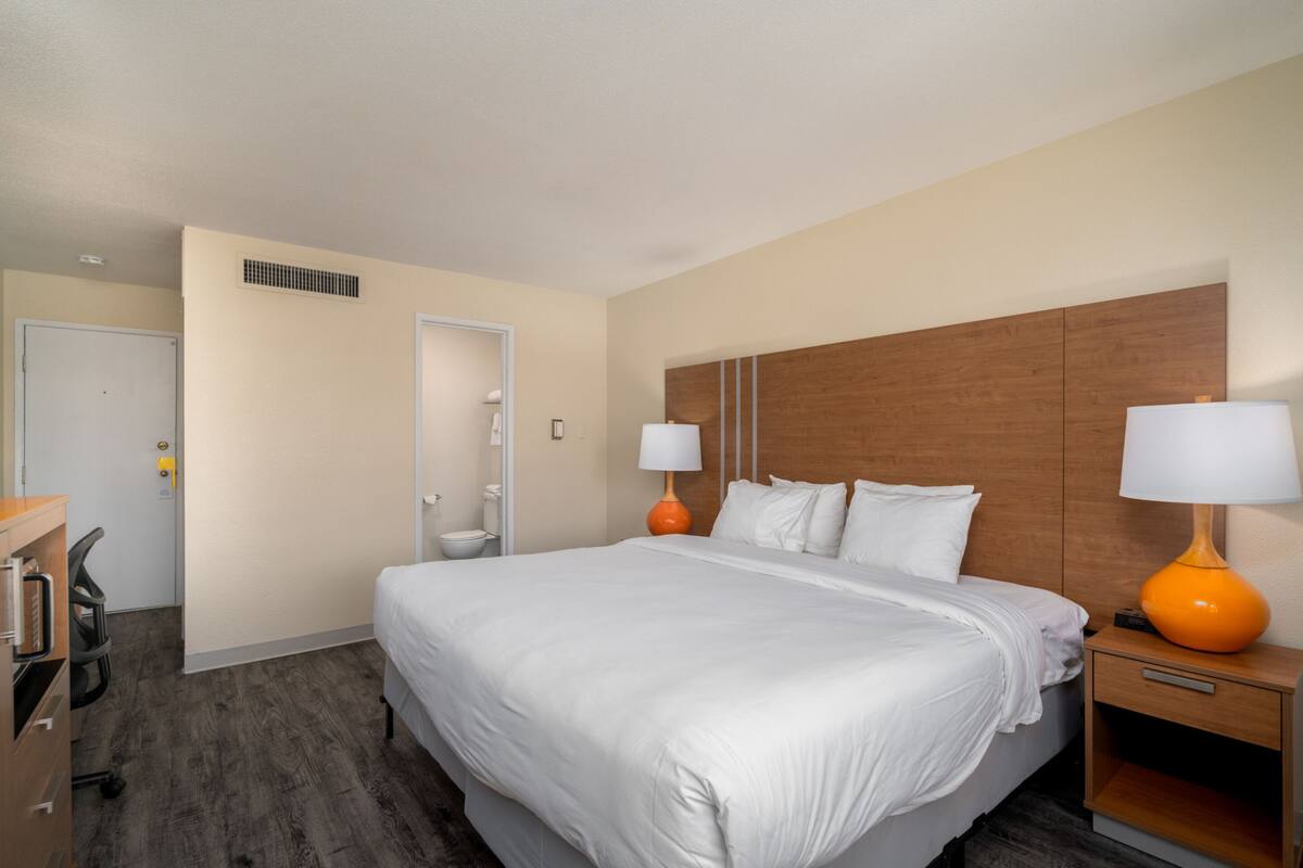 Une chambre d’hôtel avec un grand lit, deux tables de chevet avec des lampes, une tête de lit en bois, un bureau avec une chaise et une porte de salle de bain.
