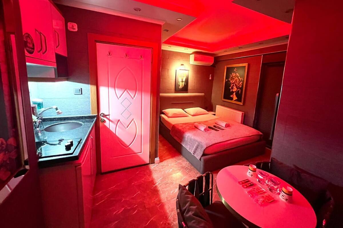 A modern hotel room with a red-lit ambiance, a bed, a small table, and a bathroom with a sink and mirror.