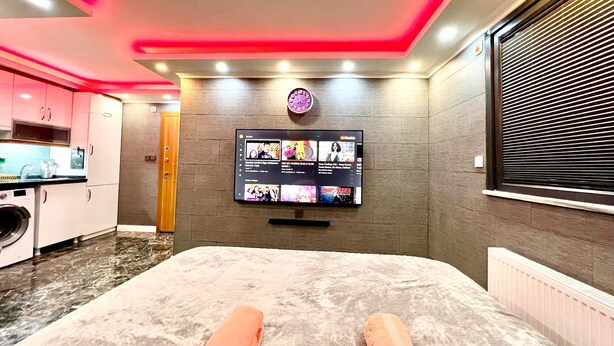 A modern hotel room with a flat-screen TV mounted on the wall, a bed with a fluffy white mattress, and a kitchen area with pink cabinets. - Levent Palace Hotel (Istanbul)