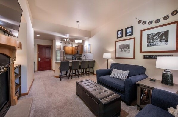 A living room with a fireplace, a sofa, a coffee table, and a dining area with chairs and a kitchen in the background.