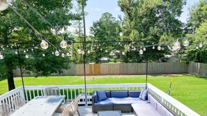 A backyard patio with a white fence, a dining table with chairs, a blue sofa, and hanging light bulbs. - Bristow Bungalow (Tupelo)