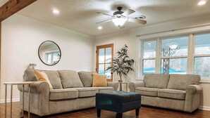 A living room with two sofas, a coffee table, a ceiling fan, a mirror, and a window with a view. - Bristow Bungalow (Tupelo)