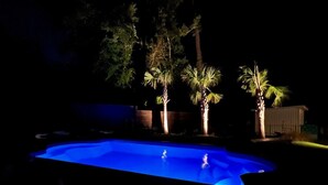 Une piscine illuminée la nuit, entourée d’arbres et bordée d’une structure en bois.