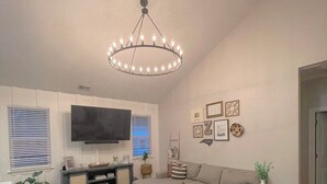 Un salon moderne avec un grand canapé d’angle gris, un téléviseur à écran plat fixé au mur et un lustre.
