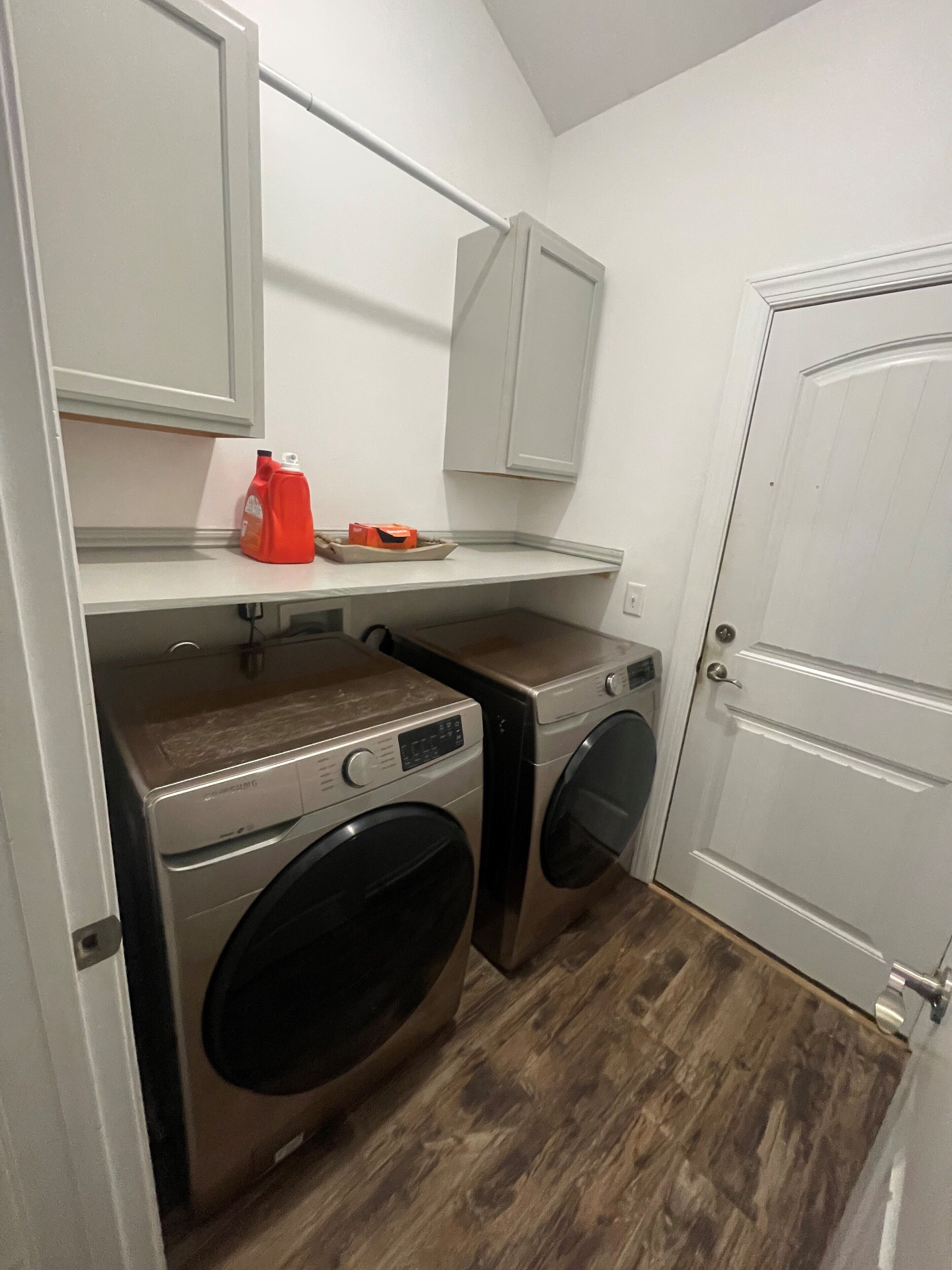 Une buanderie équipée de deux machines à laver, d’un sèche-linge, d’une étagère sur laquelle se trouve une bouteille rouge, et d’une porte blanche.
