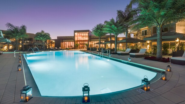 Piscina com espreguiçadeiras e palmeiras, cercada por edifícios com janelas amplas e iluminação externa.